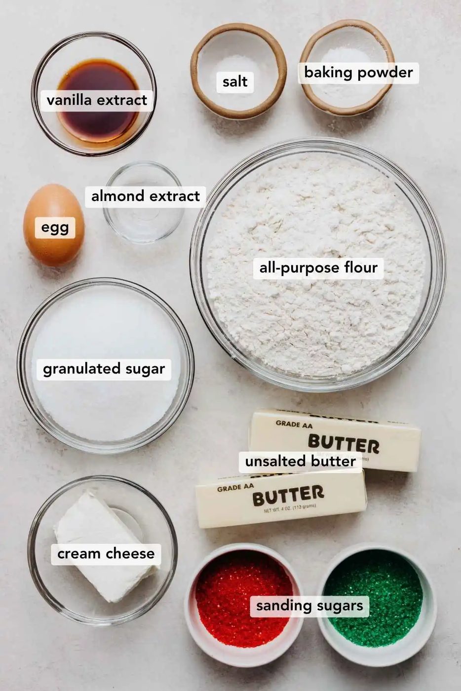 Ingredients in various size bowls labeled as vanilla extract, salt, baking powder, egg, almond extract, all purpose flour, granulated sugar, unsalted butter, cream cheese, and sanding sugar on a white marbled background.