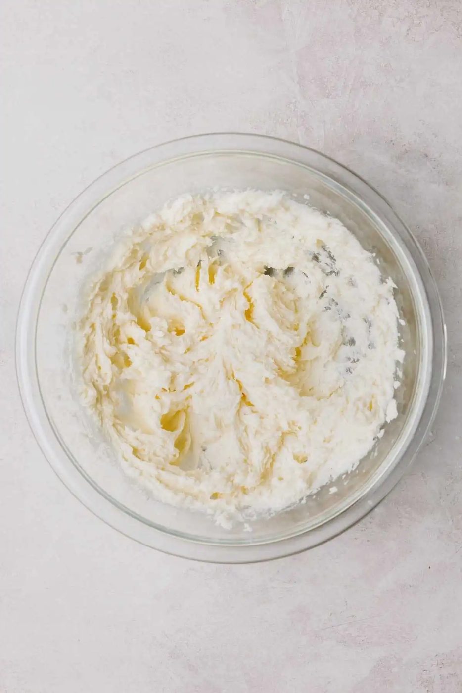 Butter, cream cheese, and granulated sugar beaten together in a large glass mixing bowl on a light grey background.