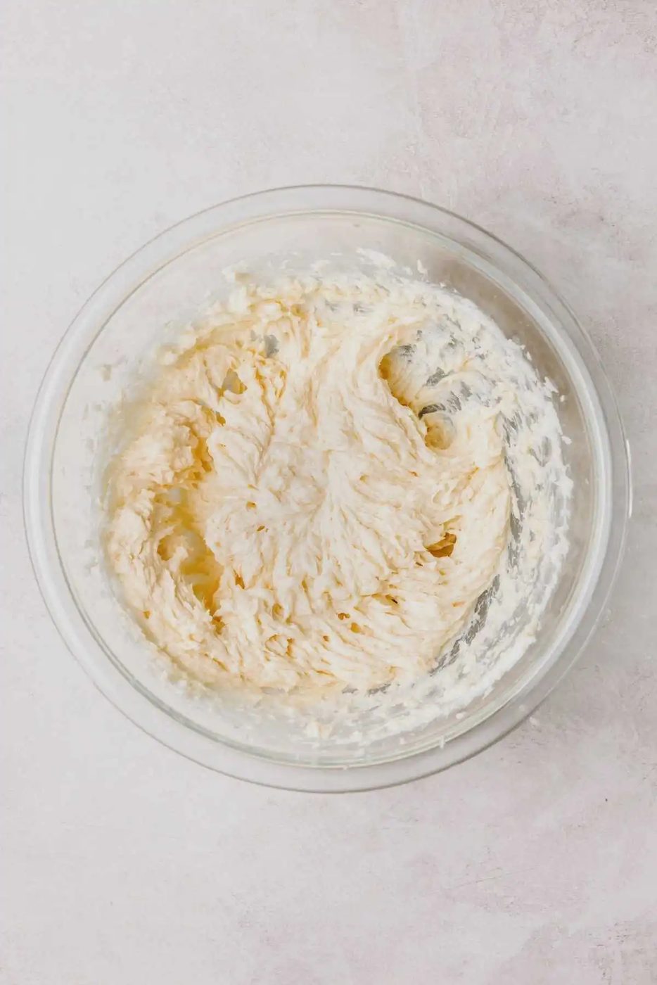 An egg, vanilla extract, and almond extract mixed into the almond cookie dough mixture in a large glass bowl on a light grey background.