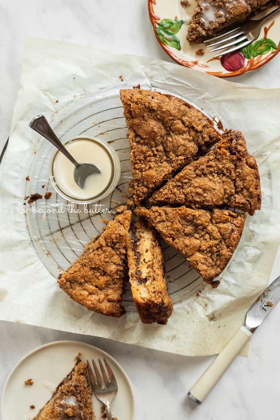 Slices of apple coffee cake on top of a parchment paper lined wire cooling rack with a small jar of simple vanilla glaze next to it | All Images © Sovuh