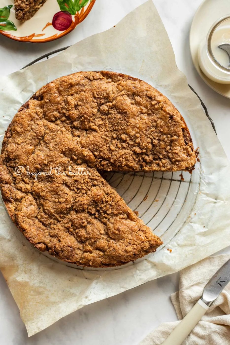 Apple coffee cake on a parchment paper lined wire cooling rack with a slice removed and a simple vanilla glaze, dessert plates, and a knife to the side | All Images © Sovuh
