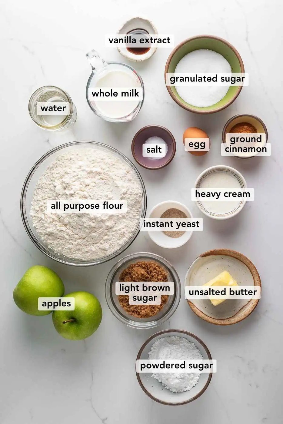 Ingredients in various size bowls labeled as vanilla extract, water, whole milk, granulated sugar, all purpose flour, salt, egg, instant yeast, heavy cream, ground cinnamon,apples, light brown sugar, butter, and powdered sugar on a white marbled background.