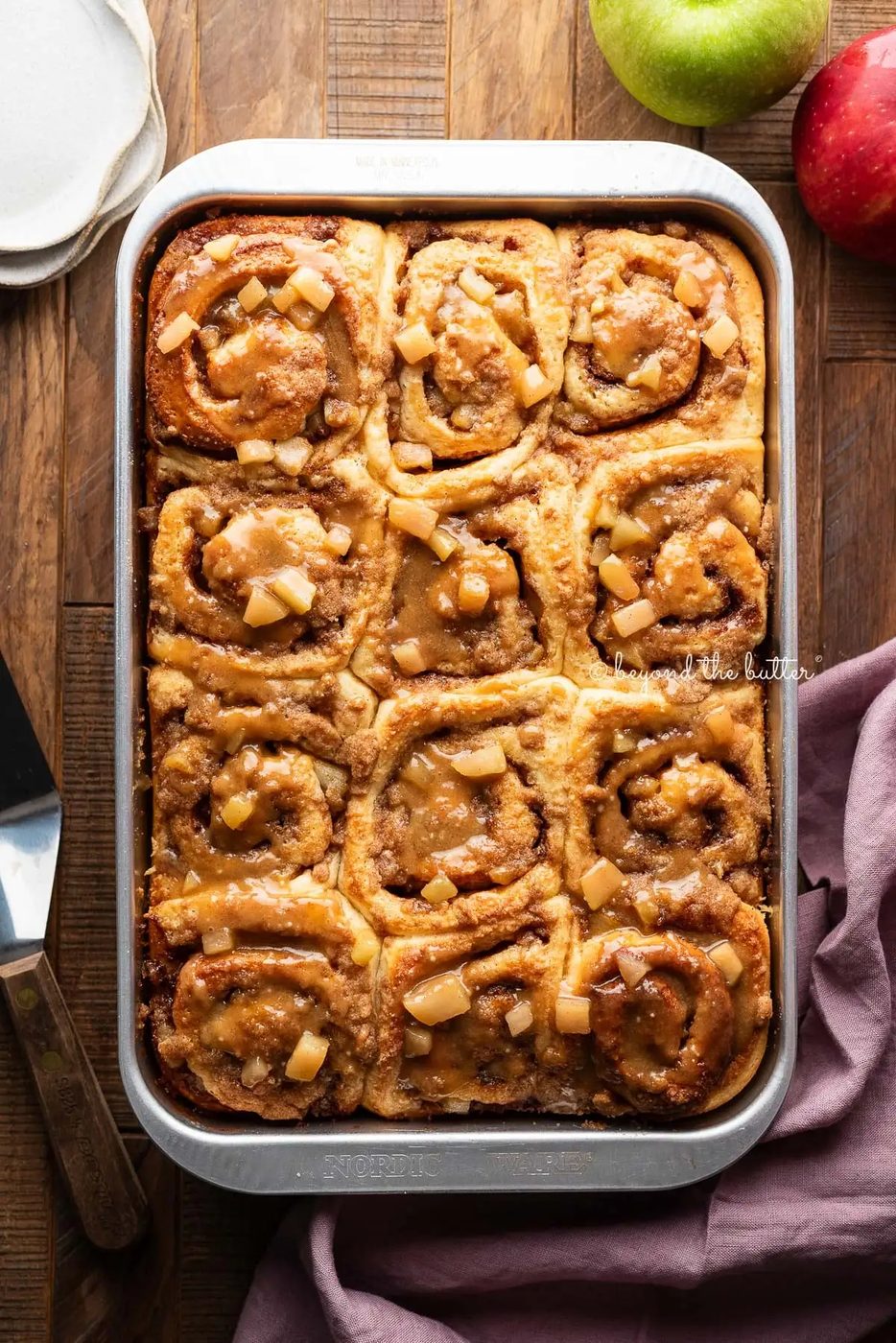Metal pan with apple streusel cinnamon rolls topped with caramel glaze placed in the center of a dark wood table with apples, ceramic dessert plates, a server spatula, and mauve colored linen napkin surrounding it.