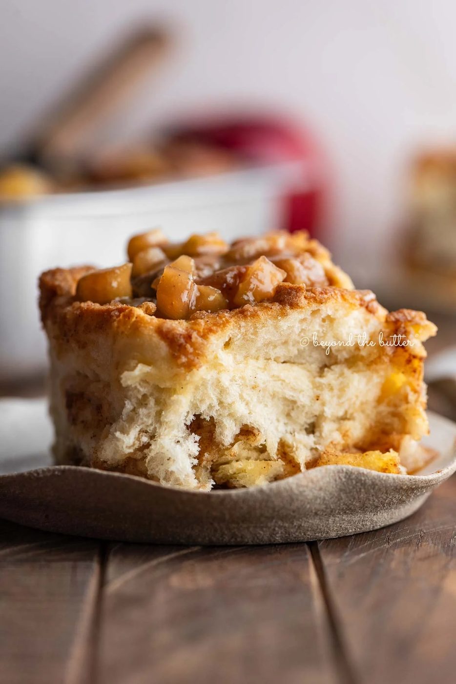 Apple streusel cinnamon roll on a ceramic dessert plate on a dark wood table with a pan of cinnamon rolls and apples in the background.