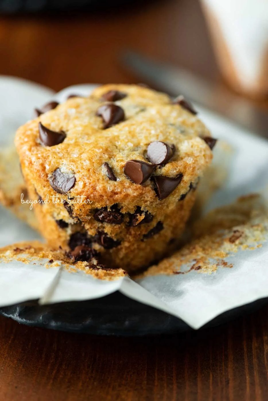 Unwrapped bakery style chocolate chip muffin on flattened muffin linerand black dessert plate | © Sovuh