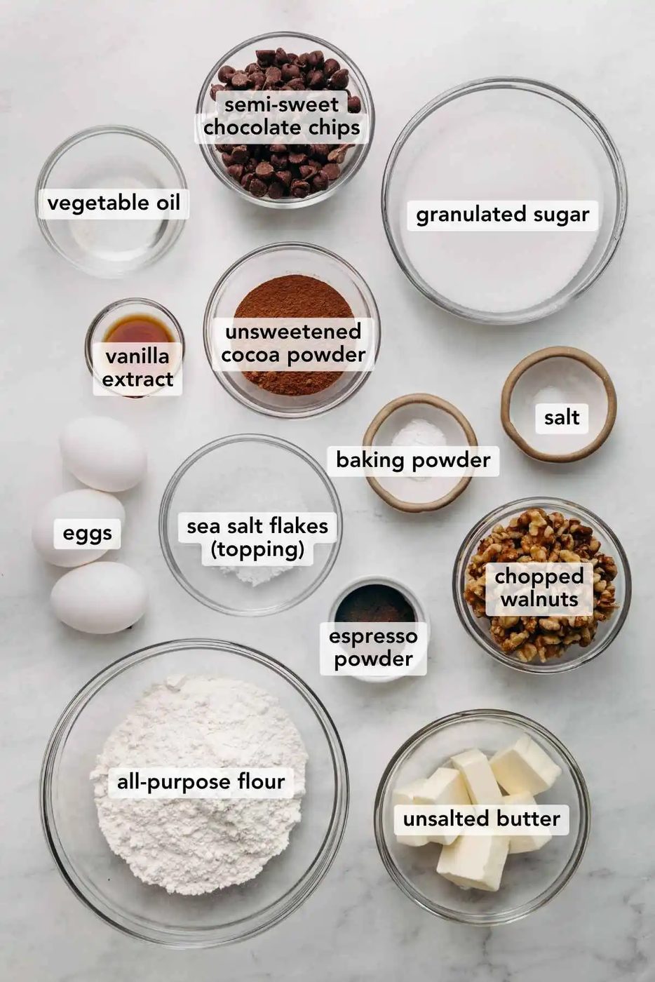 Ingredients in various size bowls labeled as vegetable oil, semi sweet chocolate chips, granulated sugar, vanilla extract, unsweetened cocoa powder, baking powder, salt, eggs, sea salt flakes (topping), chopped walnuts, all purpose flour, and unsalted butter on a white marbled background.