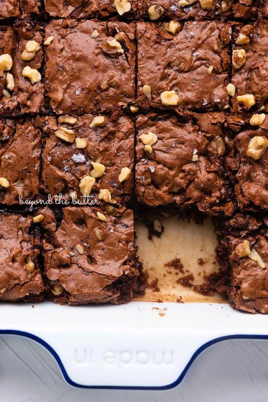 Sliced house-made fudgy brownies topped with chopped walnuts and sea salt flakes in a white and blue ceramic square baking pan lined with brown parchment paper.