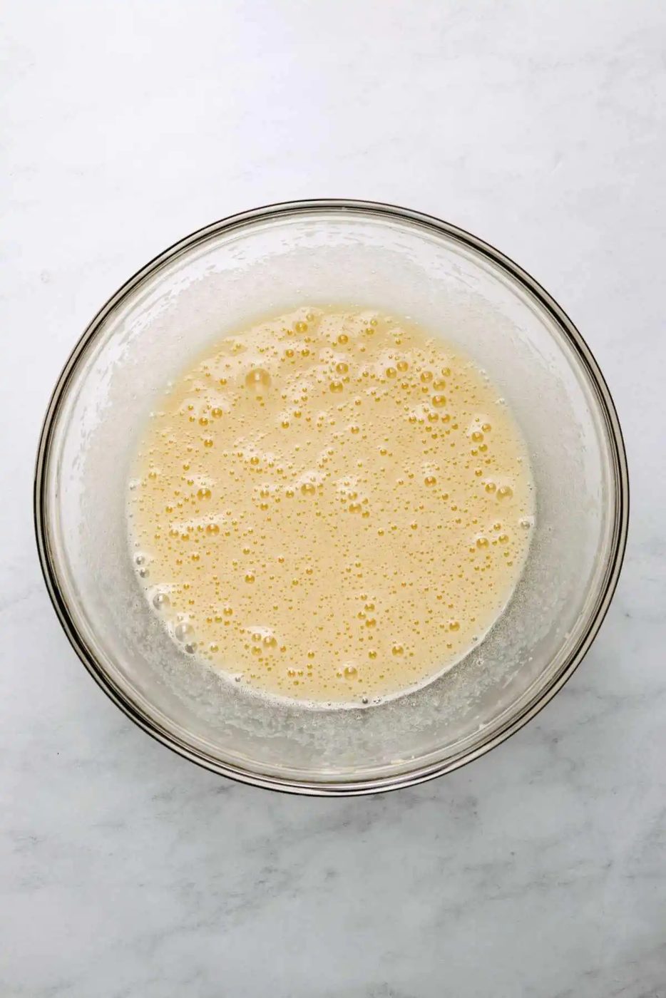 Egg, egg yolk, granulated sugar, vegetable oil, and vanilla extract whisked together in a large glass mixing bowl on a light grey background.