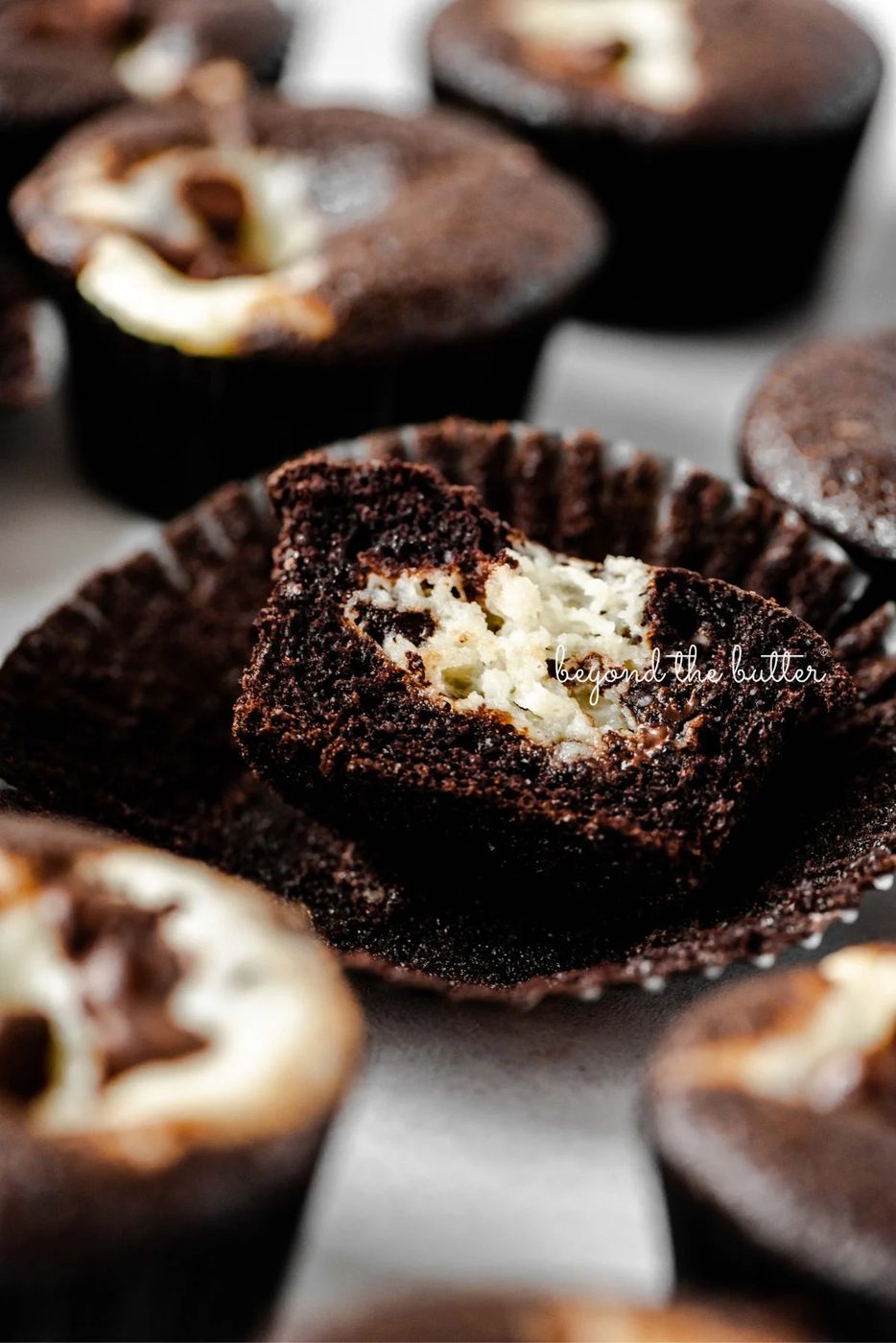 Cream cheese filled chocolate cupcakes topped with semi-sweet chocolate chips randomly placed on a light gray background with center cupcake unwrapped and sliced in half to see the cream cheese center.