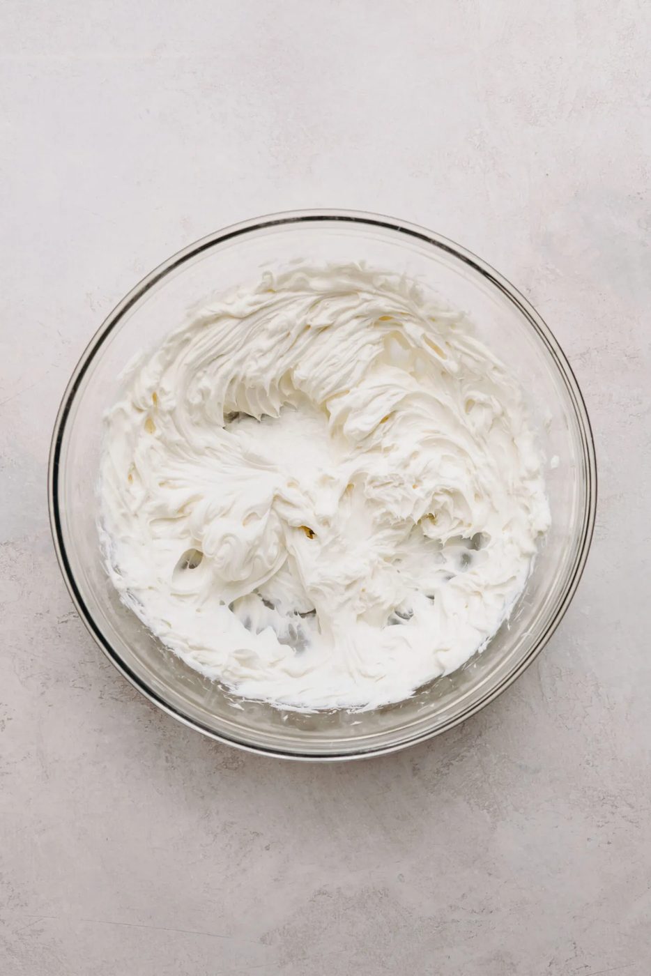 Softened and beaten cream cheese in a glass mixing bowl on a white marbled background.