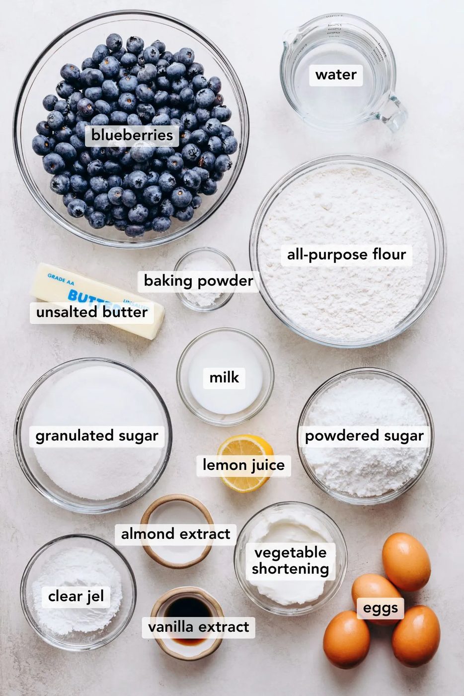 Ingredients in various size bowls and plates labeled as blueberries, water, unsalted butter, baking powder, granulated sugar, lemon juice, powdered sugar, clear jel, eggs, vanilla extract, and vegetable shortening, almond extract on a white marbled background.
