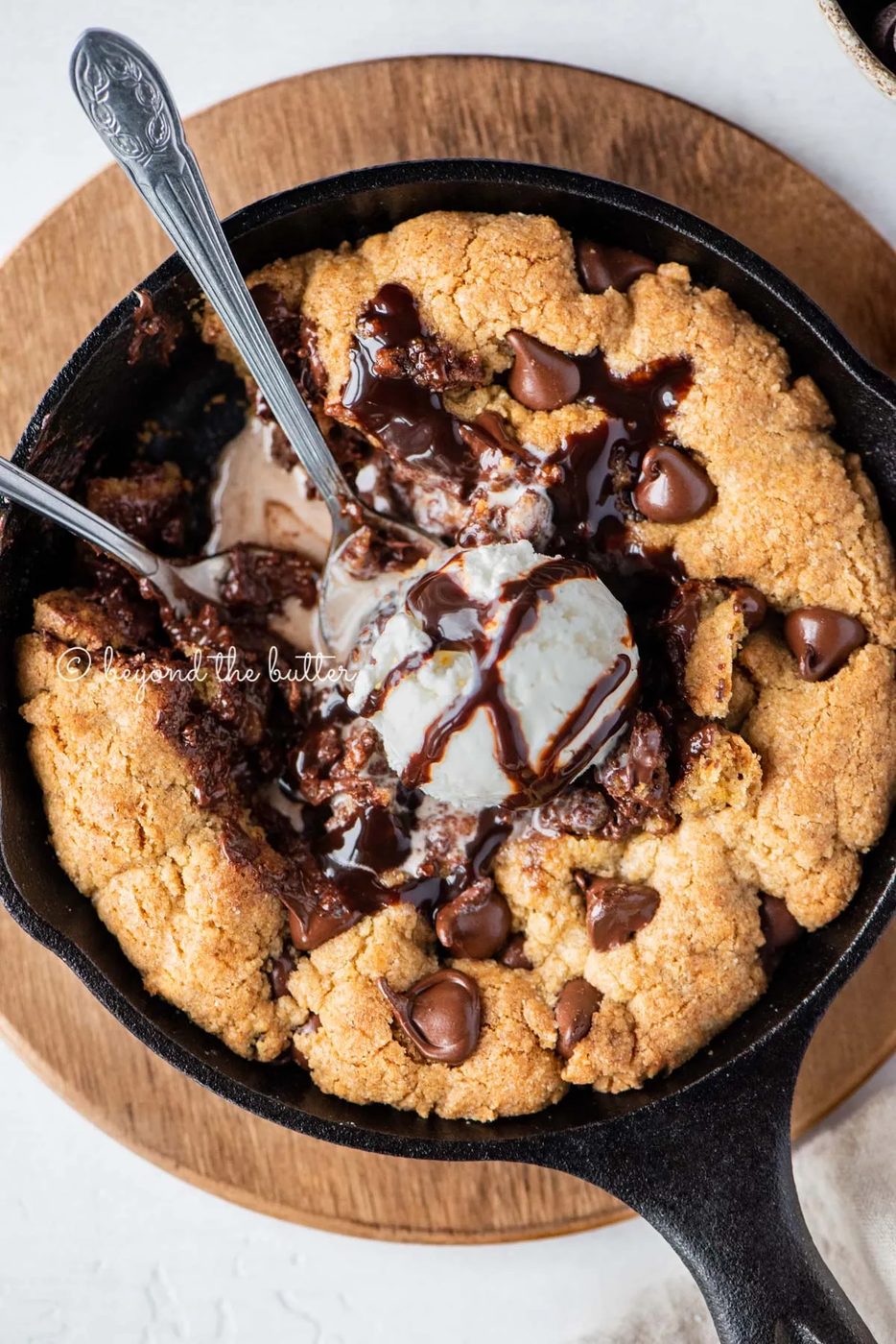 Closeup of brown butter chocolate chip skillet cookie with scoop of vanilla ice cream and two spoons resting in skillet on wood trivet | All Images © Sovuh