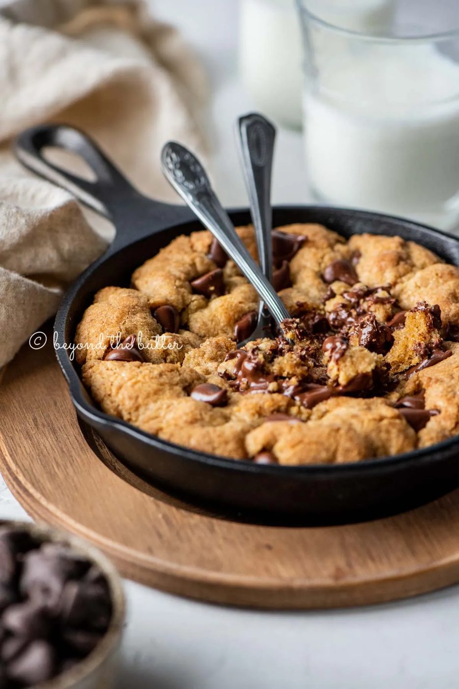 Warm brown butter chocolate chip skillet cookie with two spoons resting in skillet on wood trivet | All Images © Sovuh