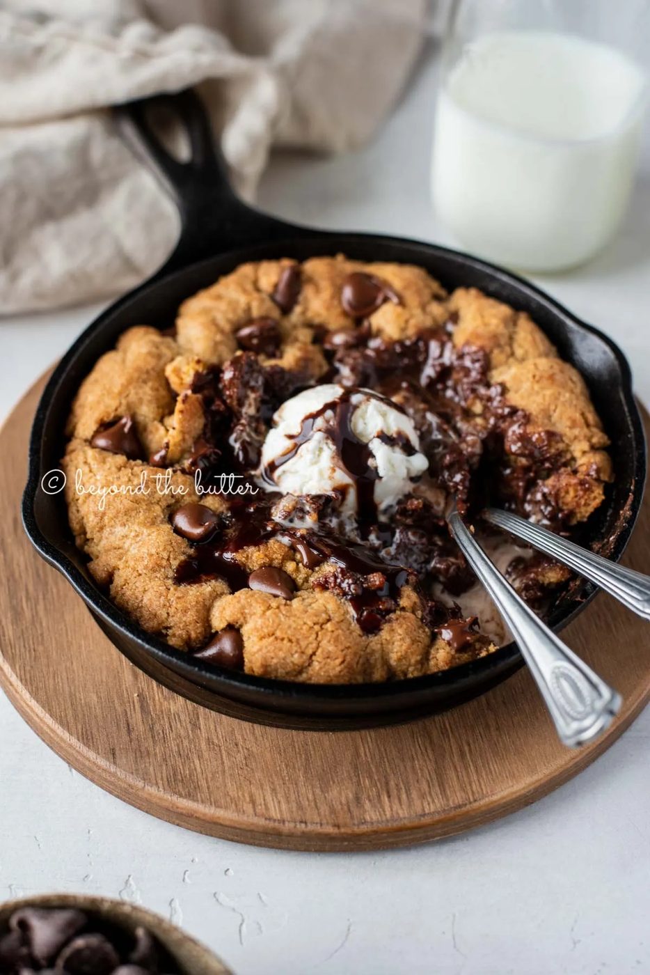 Warm chocolate chip skillet cookie with scoop of vanilla ice cream and two spoons resting in skillet on wood trivet | All Images © Sovuh