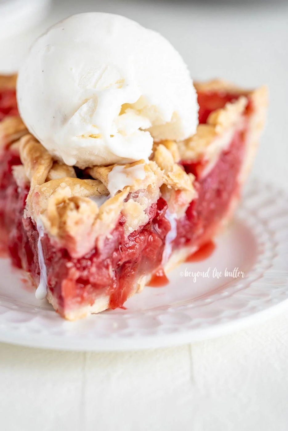 Slice of strawberry rhubarb pie with scoop of vanilla ice cream on dessert plate on white wood table.