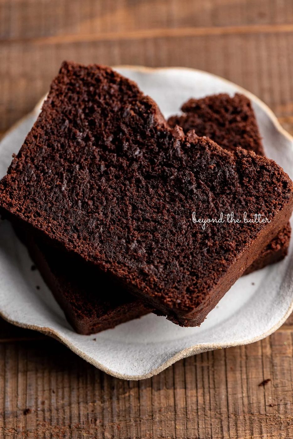Two thick slices of brownie bread on a cream colored, wavy edged ceramic dessert plate on a dark wood background.