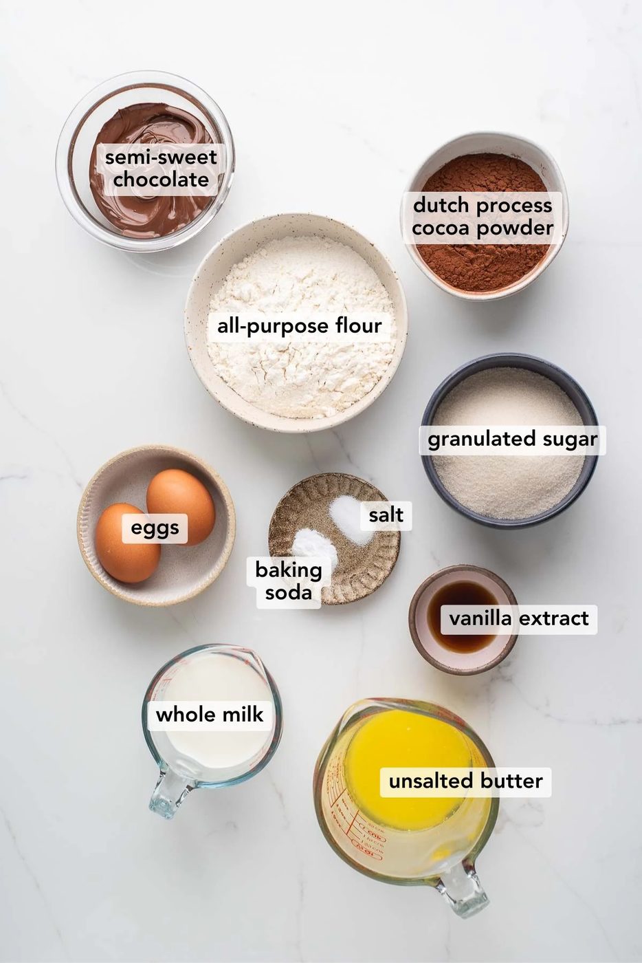 Ingredients in various size bowls and plates labeled as all semi-sweet chocolate, dutch process cocoa powder, all-purpose flour, eggs, granulated sugar, baking soda, salt, vanilla extract, whole milk, and unsalted butter on a white marbled background.