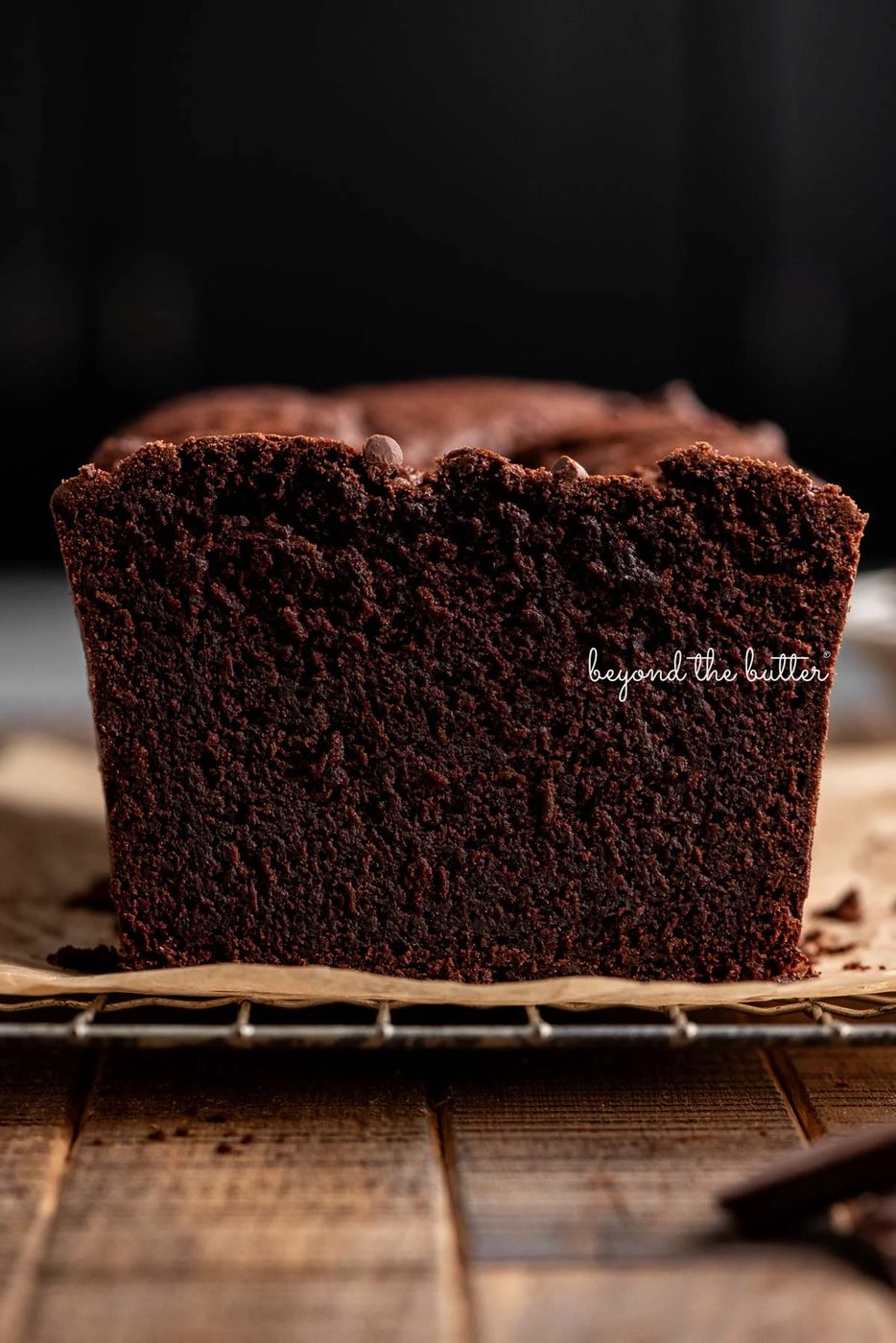 Fudgy brownie bread with end cut off on a brown parchment paper lined cooling rack on a dark wood background.