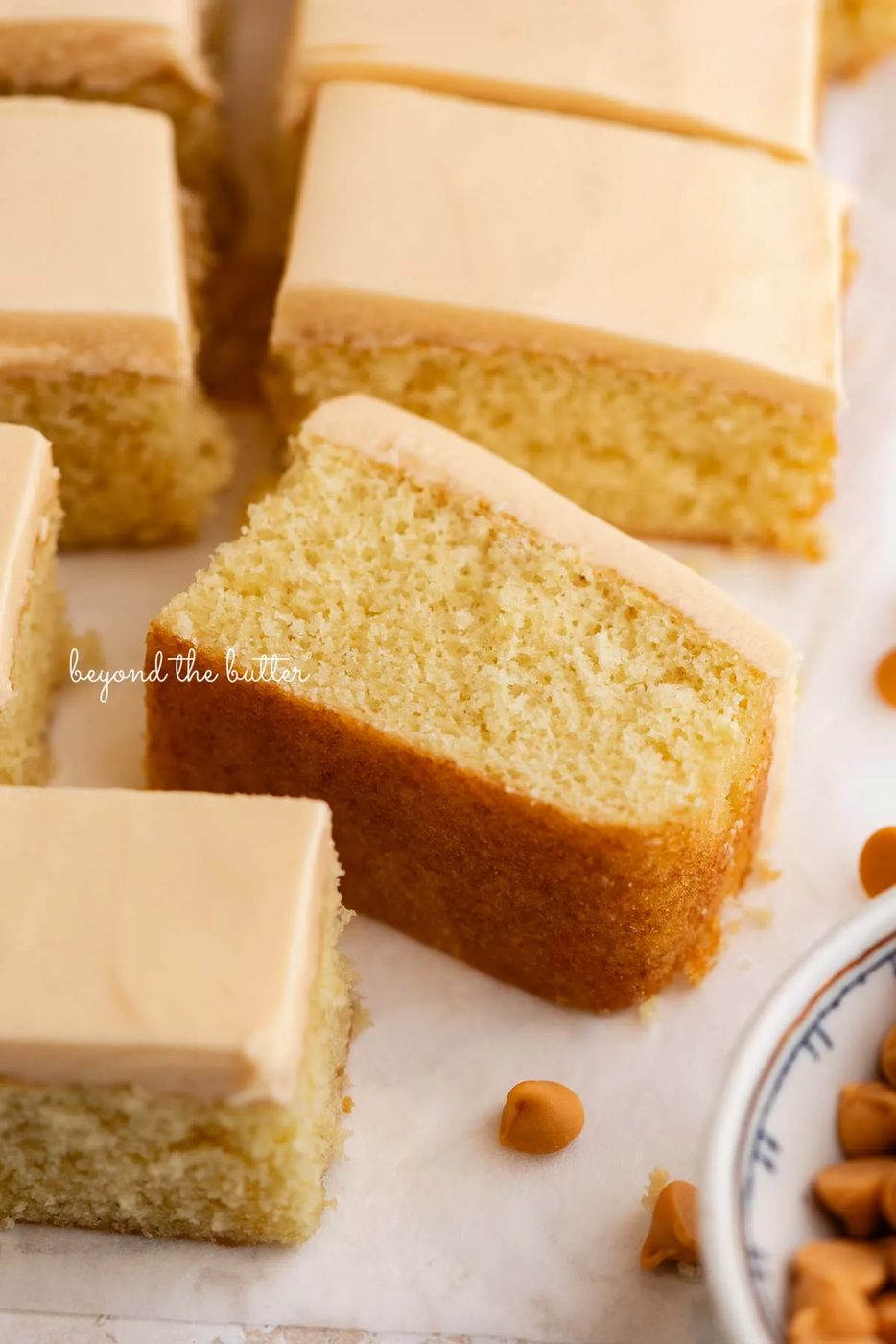 Slices of butterscotch krimpets placed on a parchment paper lined background with one slice on its side with a small decorative ceramic bowl of butterscotch chips placed next to the cake.