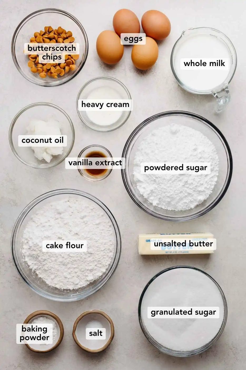 Ingredients in various size bowls labeled as butterscotch chips, eggs, whole milk, coconut oil, heavy cream, vanilla extract, powdered sugar, cake flour, unsalted butter, baking powder, salt, and granulated sugar on a white marbled background.