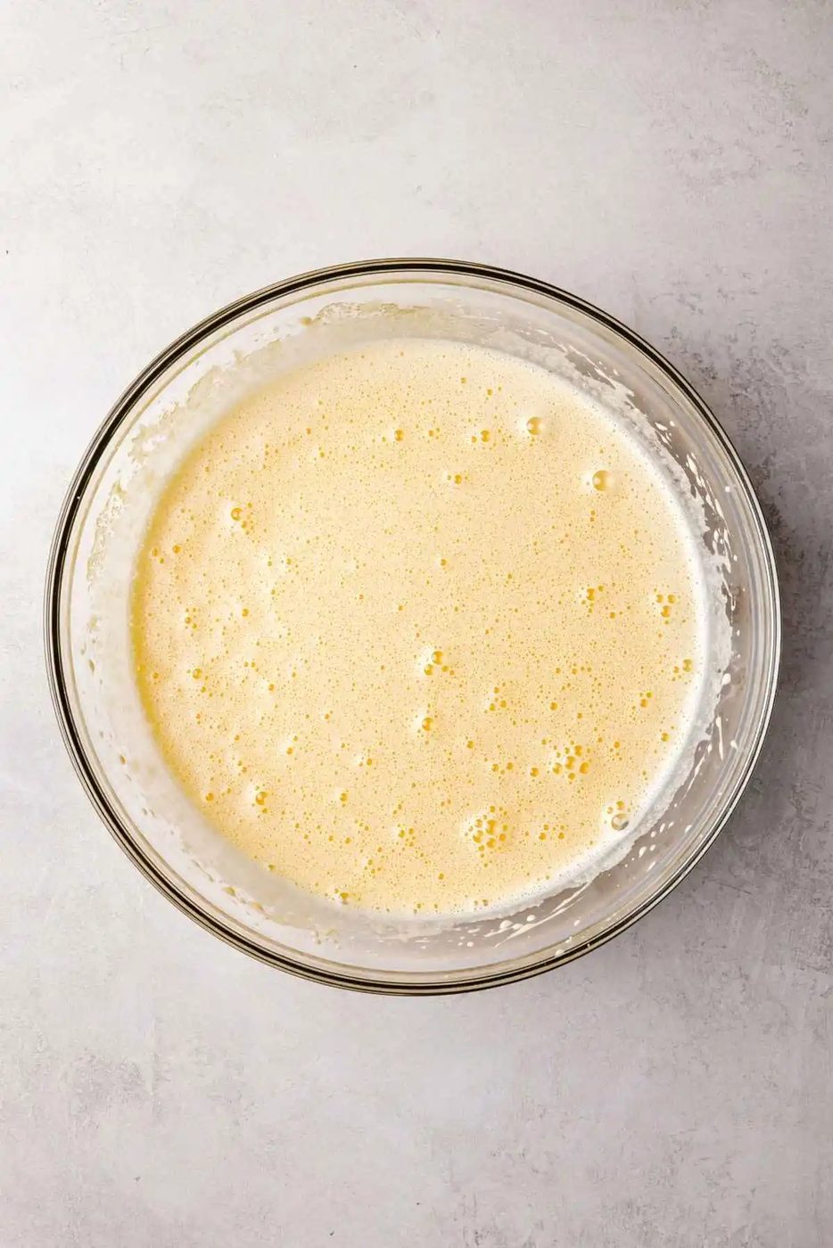 Eggs and granulated sugar whisked together until frothy and doubled in size in a large glass mixing bowl on a white and grey textured background.