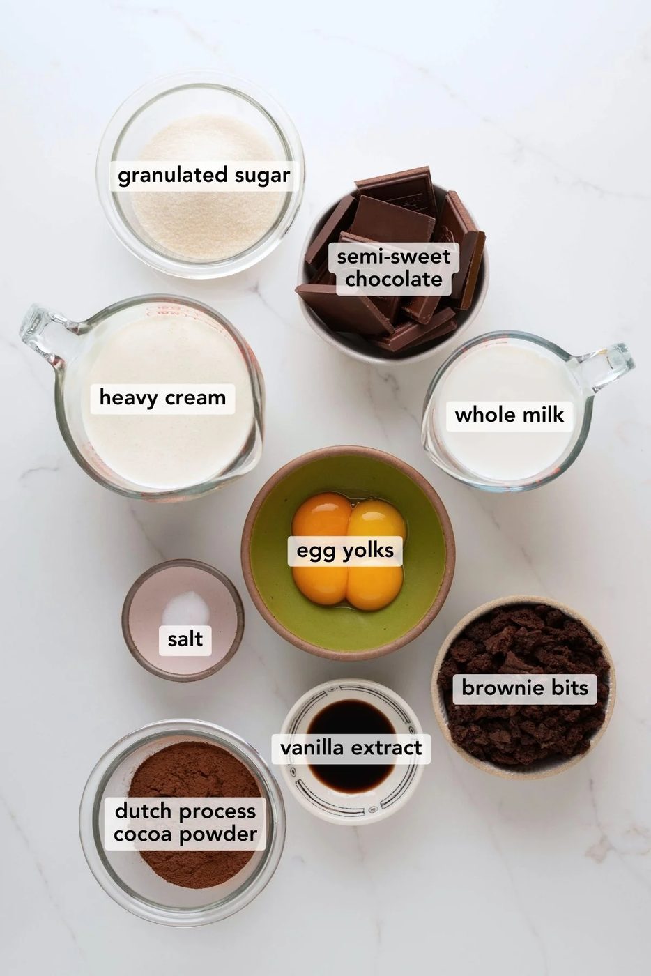 Chocolate brownie ice cream ingredients labeled and placed in separate bowl and measuring cups on a white marbled background.