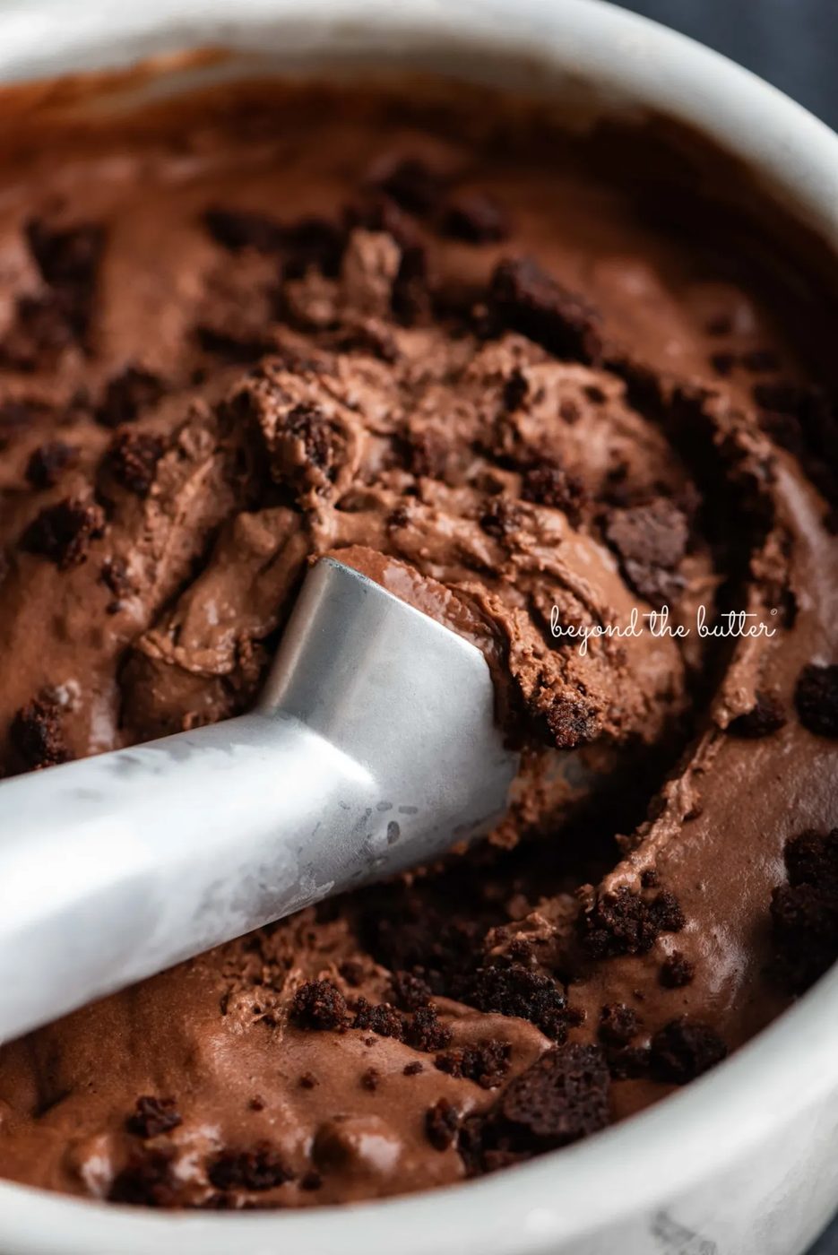 Scooping chocolate brownie ice cream out of a round stainless steel container with a chilled ice cream scoop.