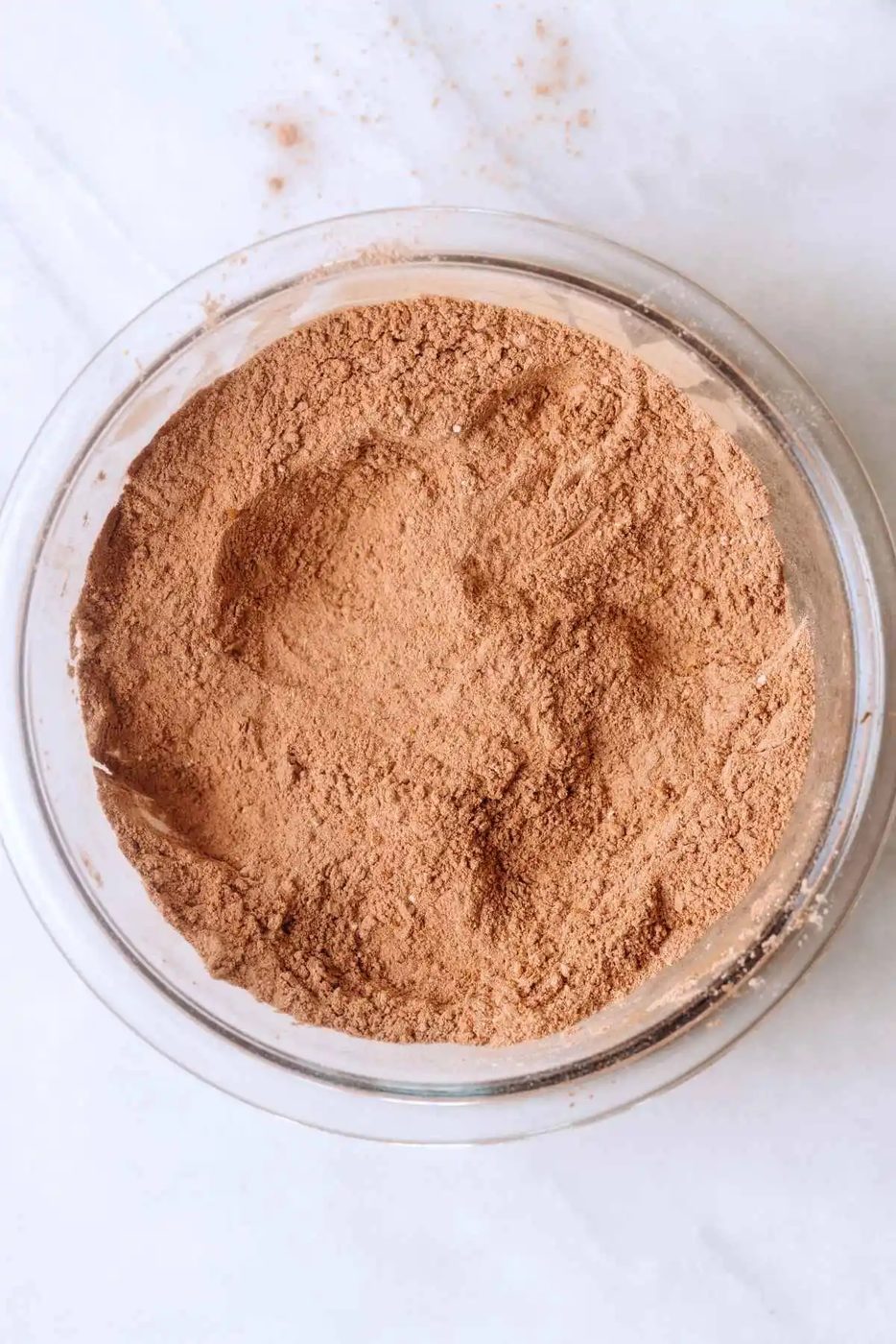 Whisked all purpose flour, baking soda, baking powder, and unsweetened cocoa powder in a large glass mixing bowl on a white and beige marbled background.