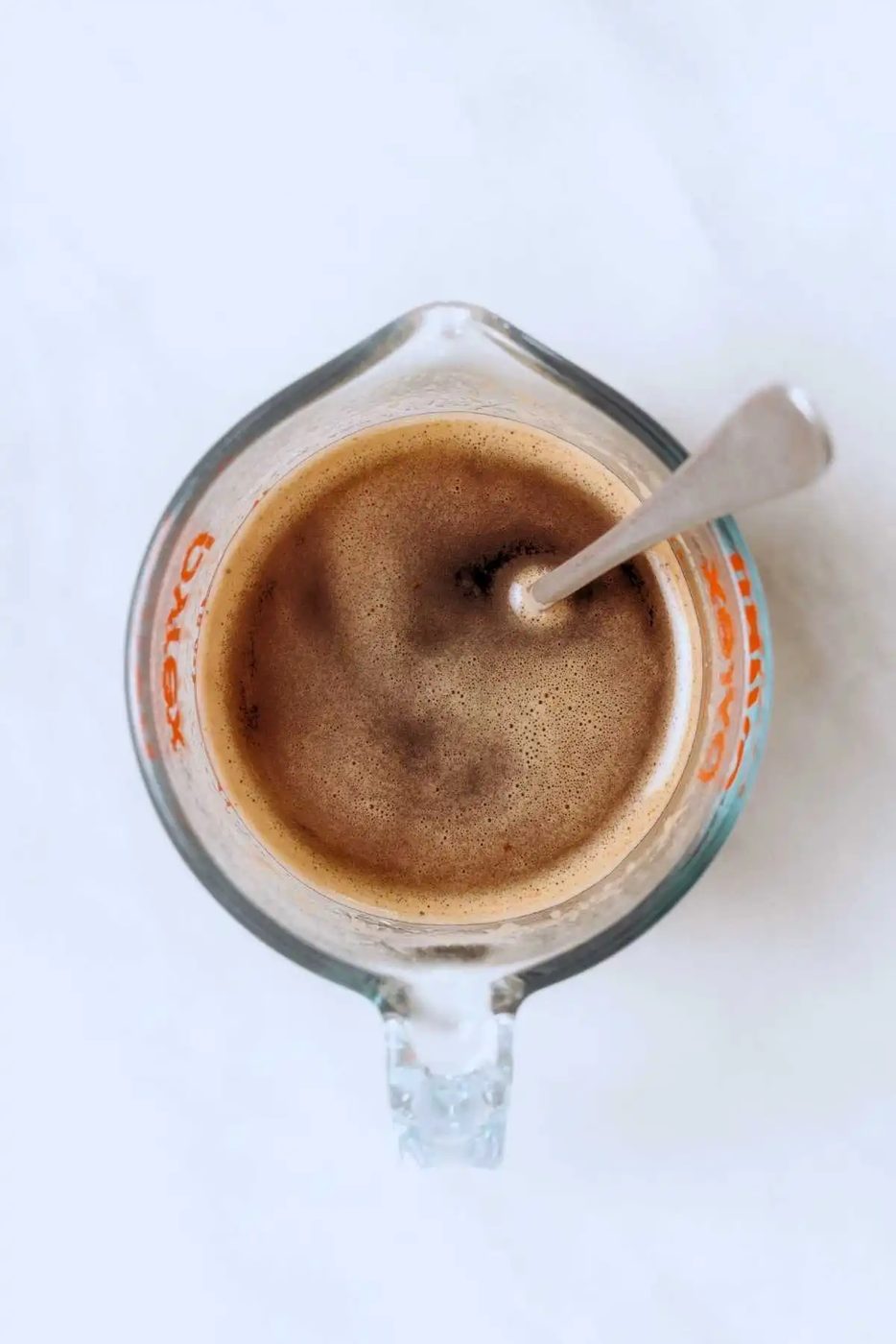 Espresso powder and boiling water stirred together in a glass measuring cup with a spoon on a white background.