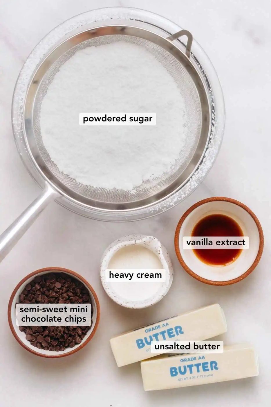 Ingredients in various size bowls labeled as powdered sugar, semi sweet mini chocolate chips, vanilla extract, heavy cream, and unsalted butter on a white marbled background.