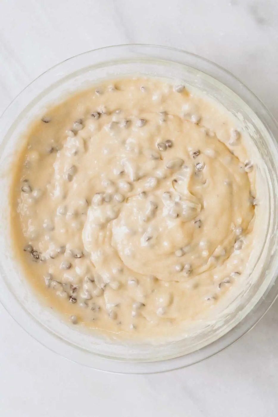 Chocolate chip cake batter in a large glass mixing bowl on a white marbled background.
