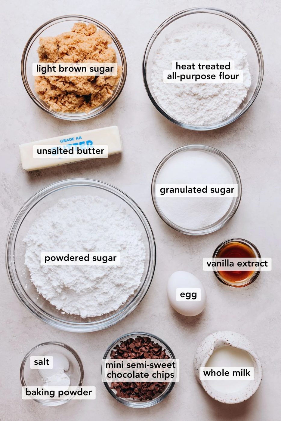 Ingredients in various size bowls and plates labeled as light brown sugar, heat treated all purpose flour, unsalted butter, granulated sugar, powdered sugar, vanilla extract, egg, salt, baking powder, mini semi-sweet chocolate chips, and whole milk on a white marbled background.
