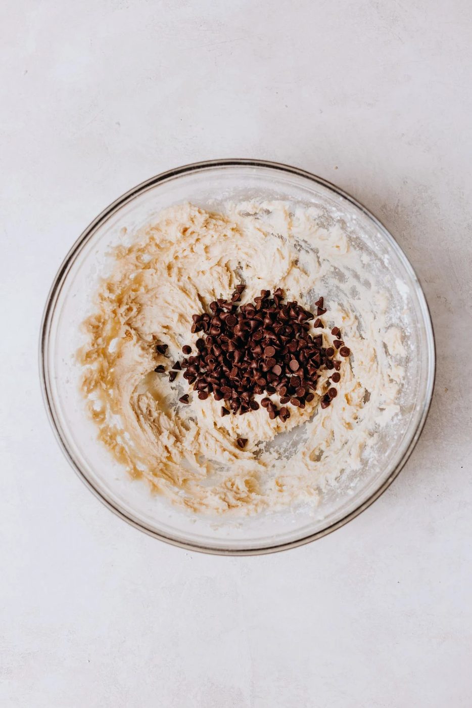 Folding mini chocolate chips into edible chocolate chip cookie dough mixture in a glass mixing bowl on a white marbled background.
