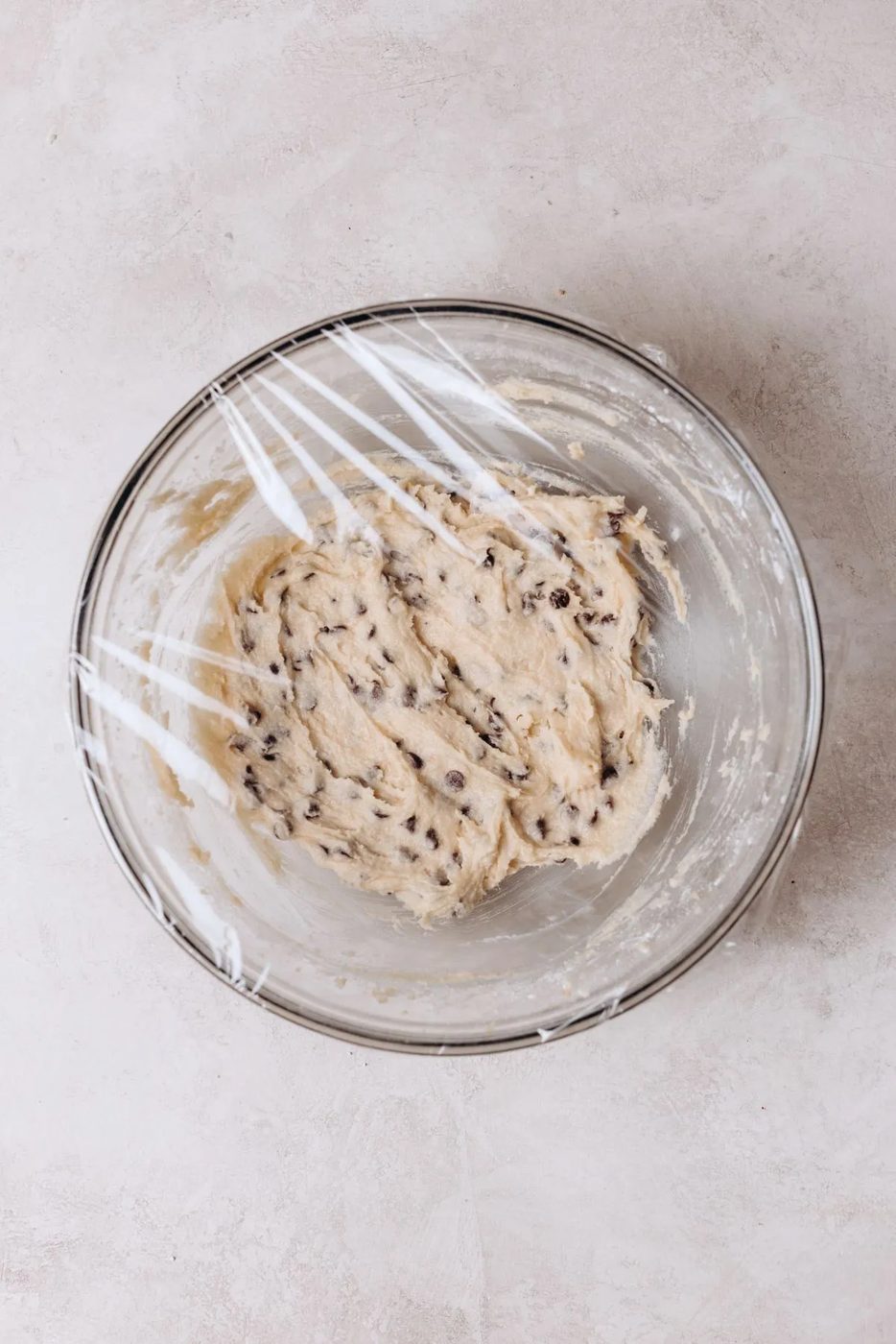Glass mixing bowl with edible chocolate chip cookie dough covered in plastic wrap on a white marbled background.
