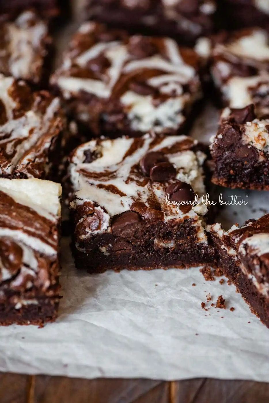 Sliced chocolate chip cream cheese brownies on white parchment paper lined placed on a dark wood background.