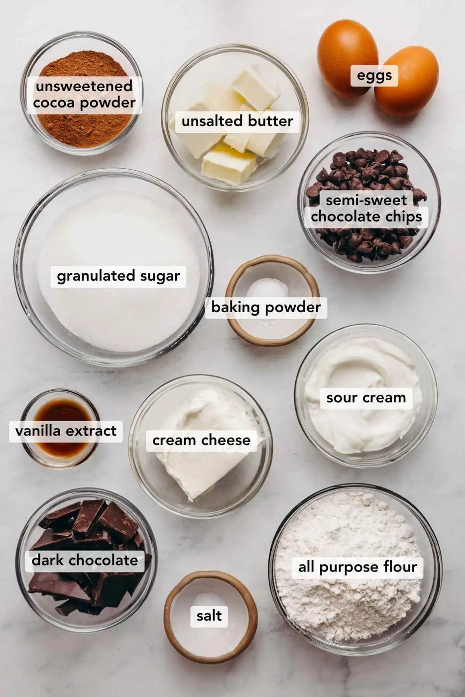 Ingredients in various size bowls labeled as unsweetened cocoa powder, unsalted butter eggs, granulated sugar, baking powder, semi sweet chocolate chips, vanilla extract, cream cheese, sour cream, dark chocolate, salt, and all purpose flour on a white marbled background.