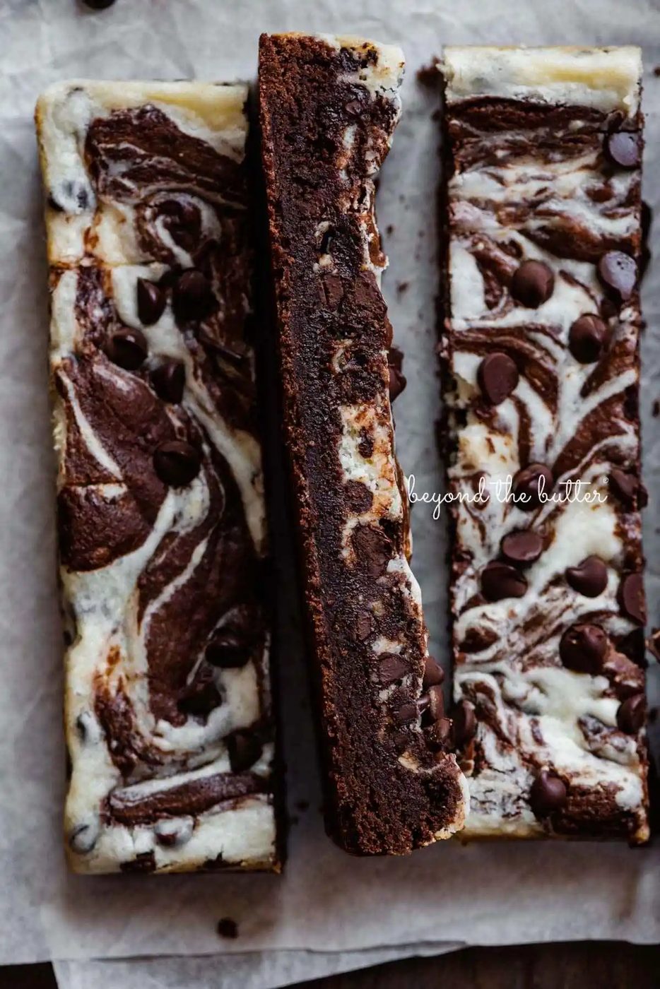 Chocolate chip cream cheese brownies cut into vertical bars on white parchment paper.
