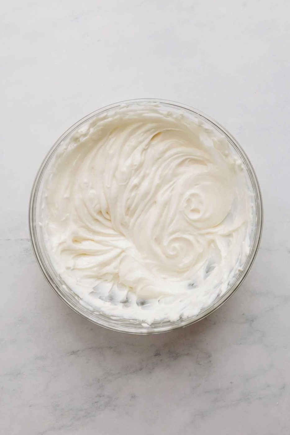 Cream cheese, sour cream, and sugar mixed together in a large glass mixing bowl on a white and grey marbled background.