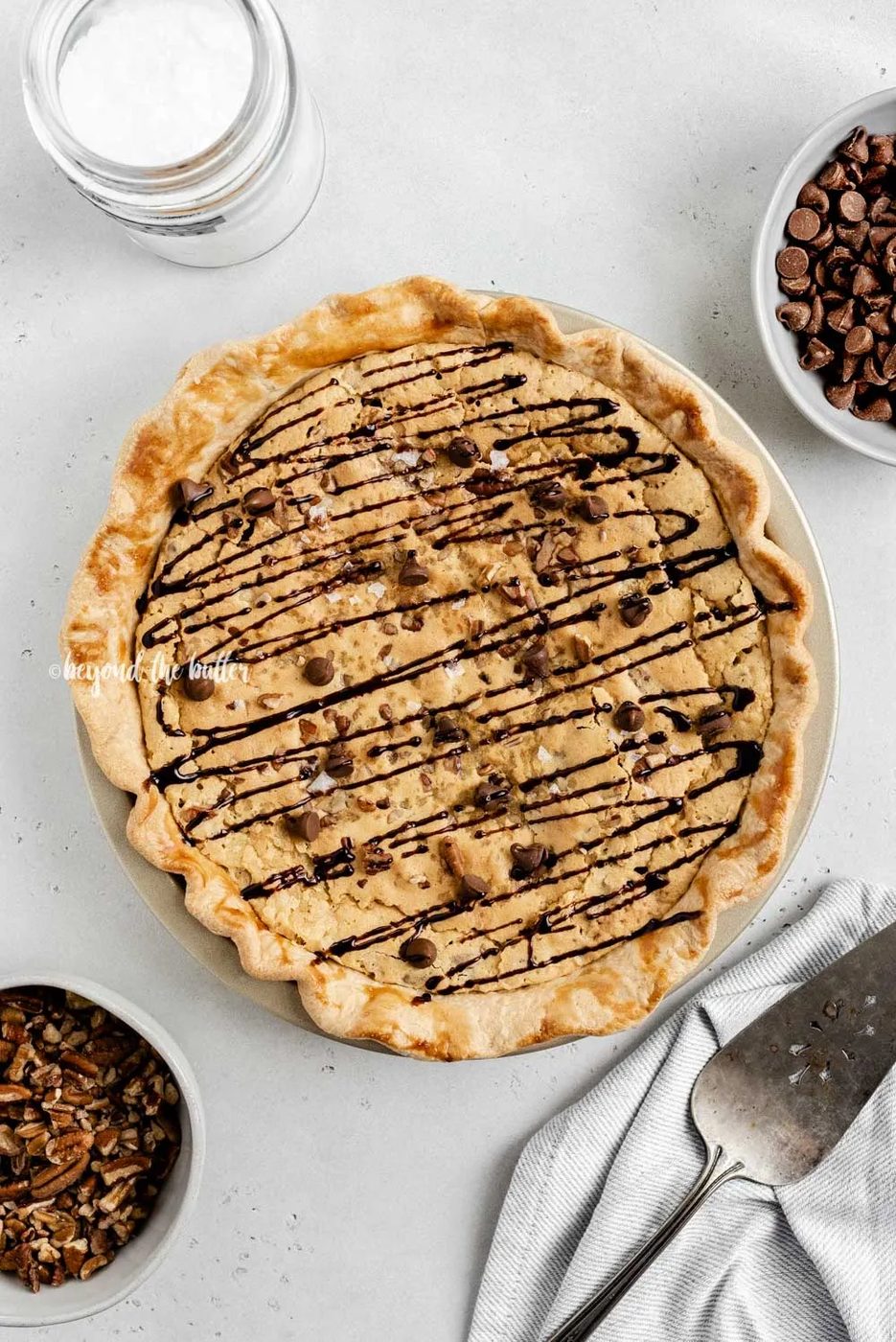 Overhead image of chocolate chip pie drizzled with chocolate syrup with bowls of chocolate chips, pecans, and a server | All Images © Sovuh