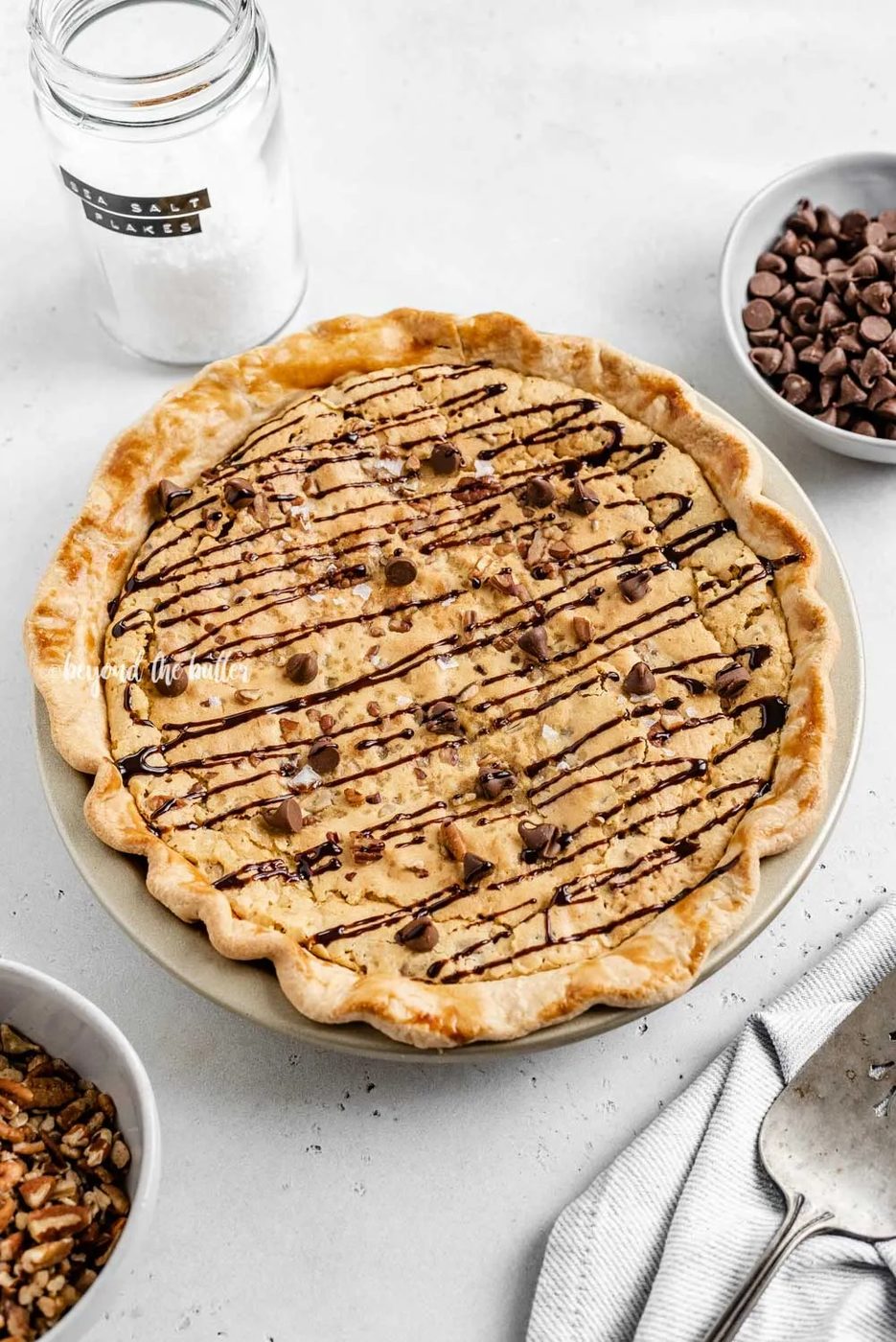 Overhead angled image of chocolate chip pie drizzled with chocolate syrup with bowls of chocolate chips, pecans, and a server | All Images © Sovuh