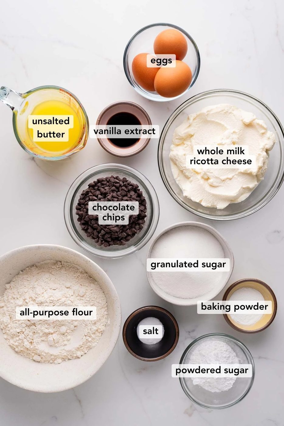Ingredients in various size bowls labeled as semi-sweet chocolate chips, whole milk ricotta cheese, unsalted butter, granulated sugar, baking powder, eggs, salt, vanilla extract, powdered sugar, and all-purpose flour on a white marbled background.