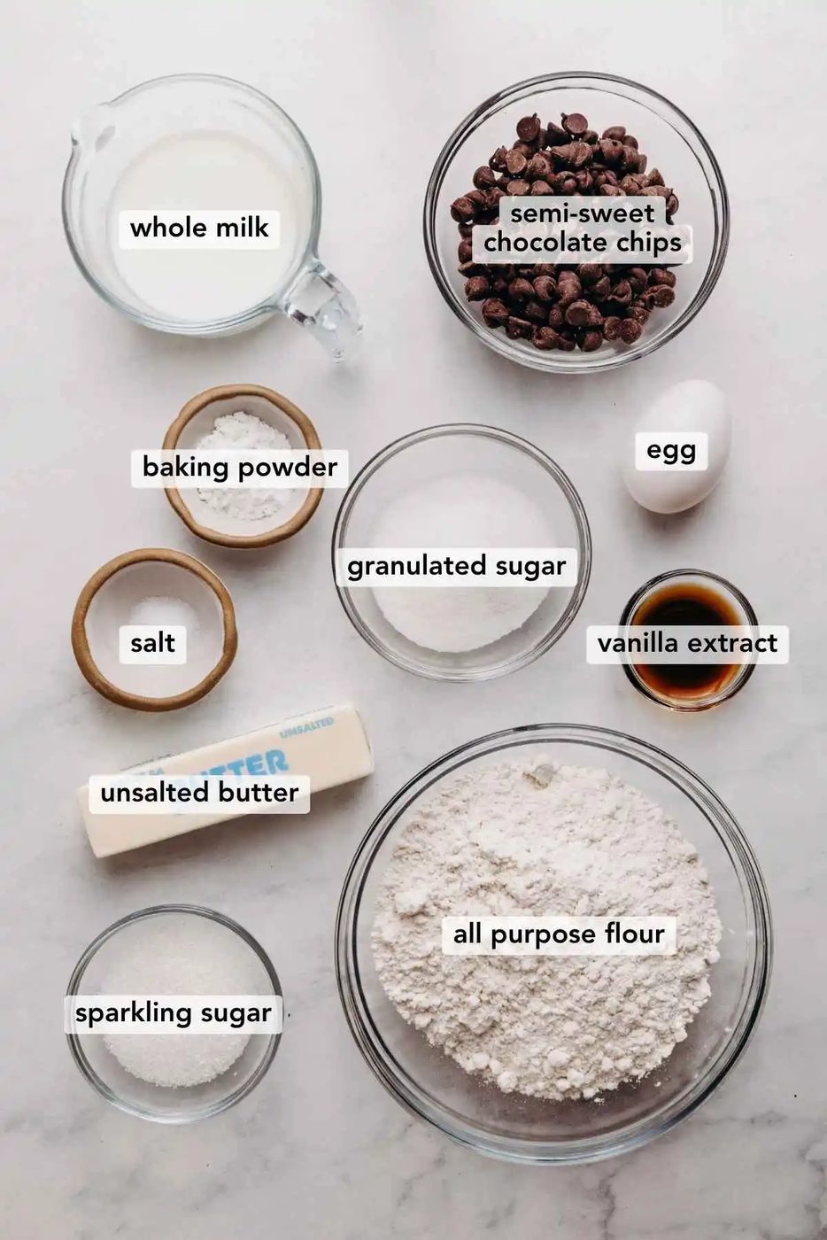 Ingredients in various size bowls labeled as whole milk, semi sweet chocolate chips, baking powder, granulated sugar, egg, salt, vanilla extract, unsalted butter, sparkling sugar, and all purpose flour on a white marbled background.