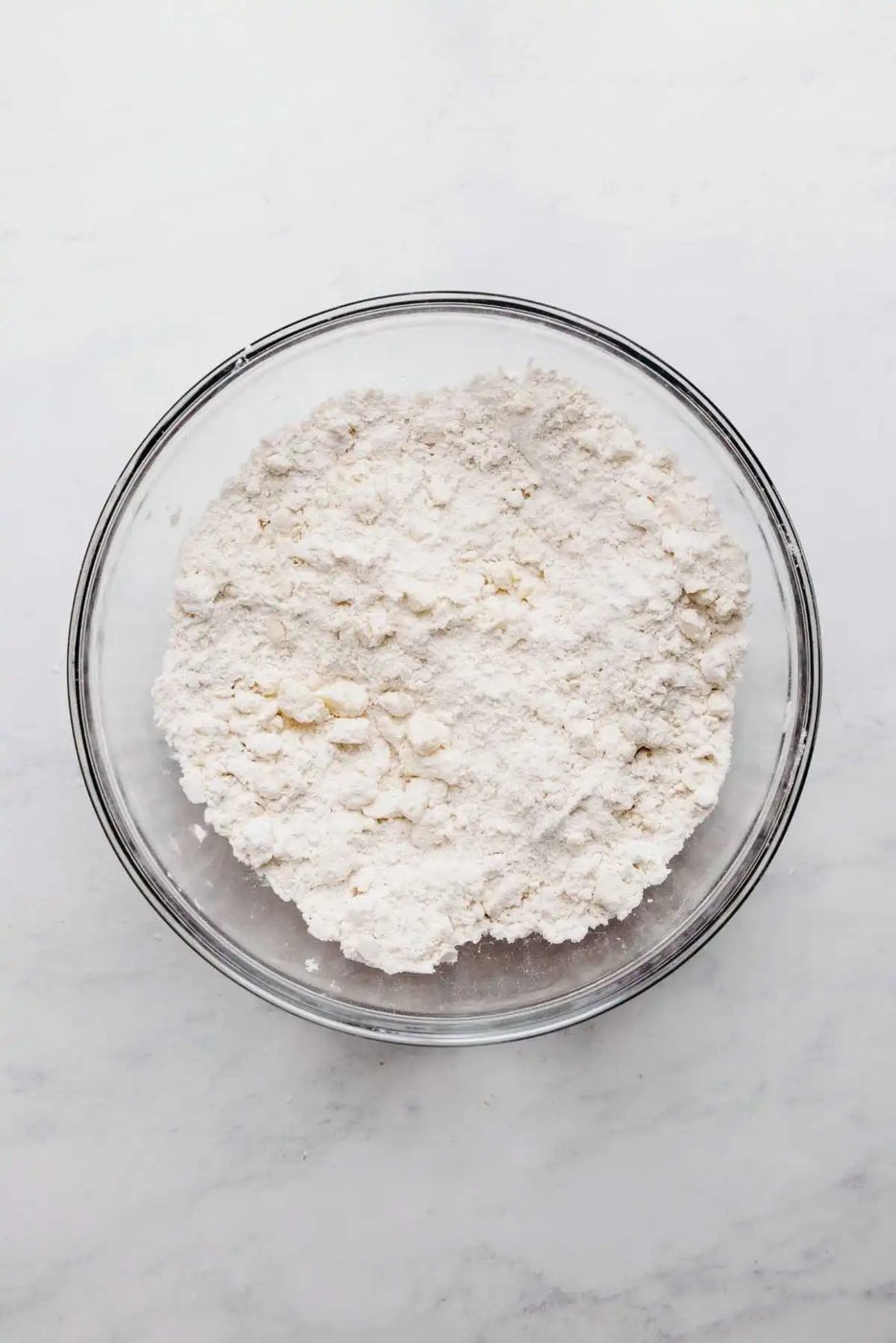 Butter cut into flour mixture in large glass mixing bowl on a white and grey marbled background.
