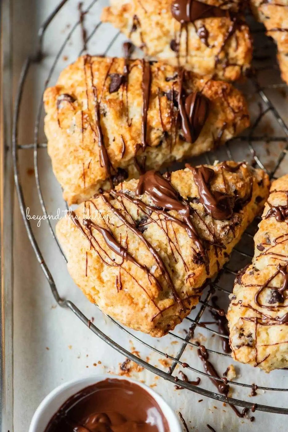 Chocolate chip scones drizzled with melted chocolate on a round wire cooling rack with a small bowl of melted chocolate placed close by.