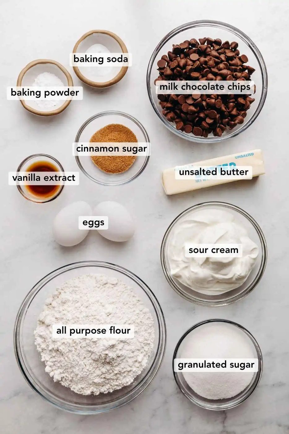 Ingredients in various size bowls labeled as baking soda, baking powder, milk chocolate chips, vanilla extract, cinnamon sugar, unsalted butter, eggs, sour cream, all purpose flour, and granulated sugar on a white marbled background.