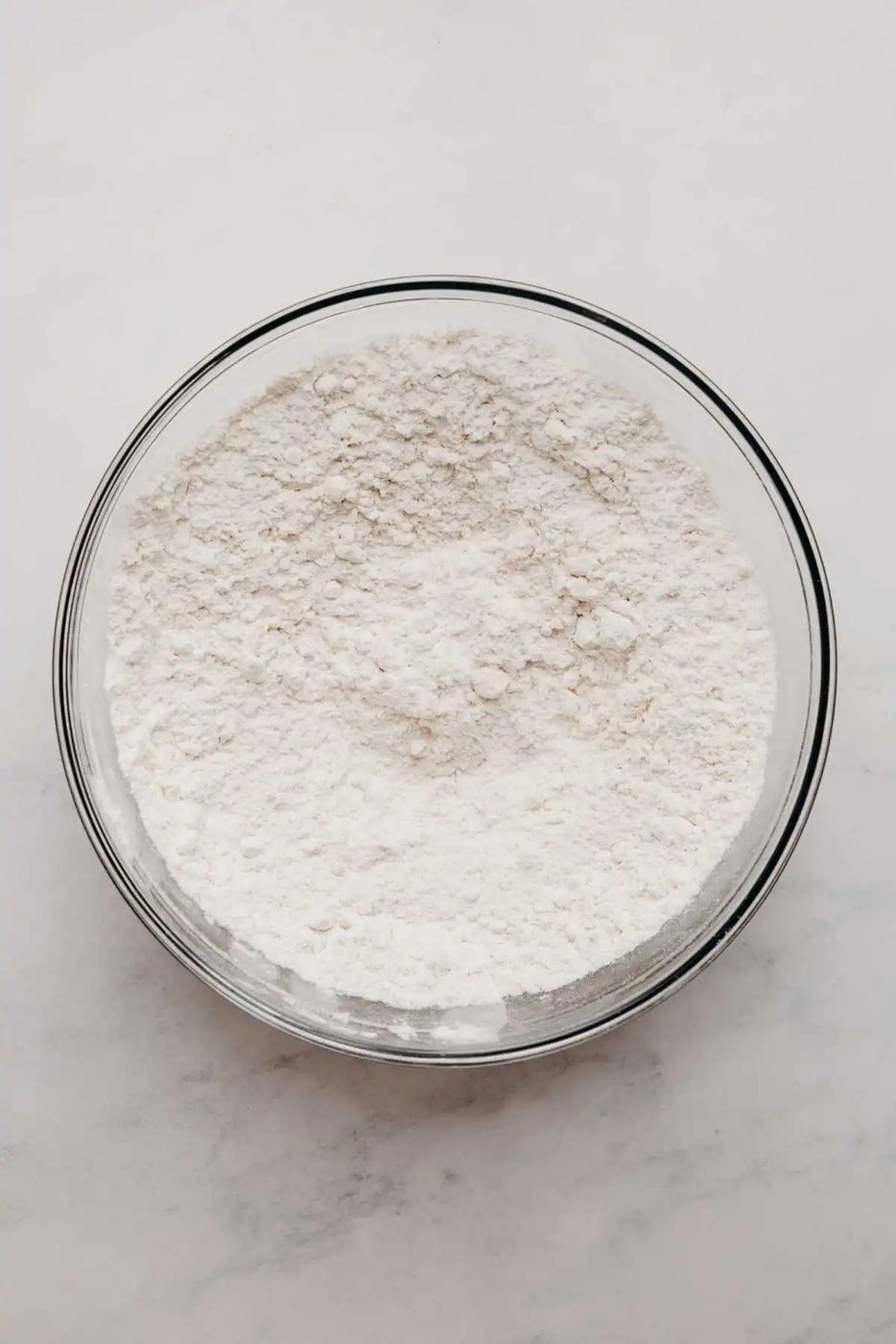 All purpose flour, baking powder, and baking soda whisked together in a large glass mixing bowl on a white marbled background.