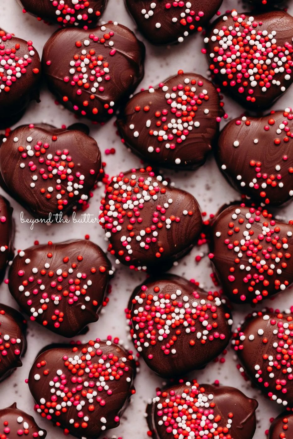 Chocolate covered cookie dough hearts topped with red, pink, and white sprinkles placed on a white parchment paper lined baking sheet.