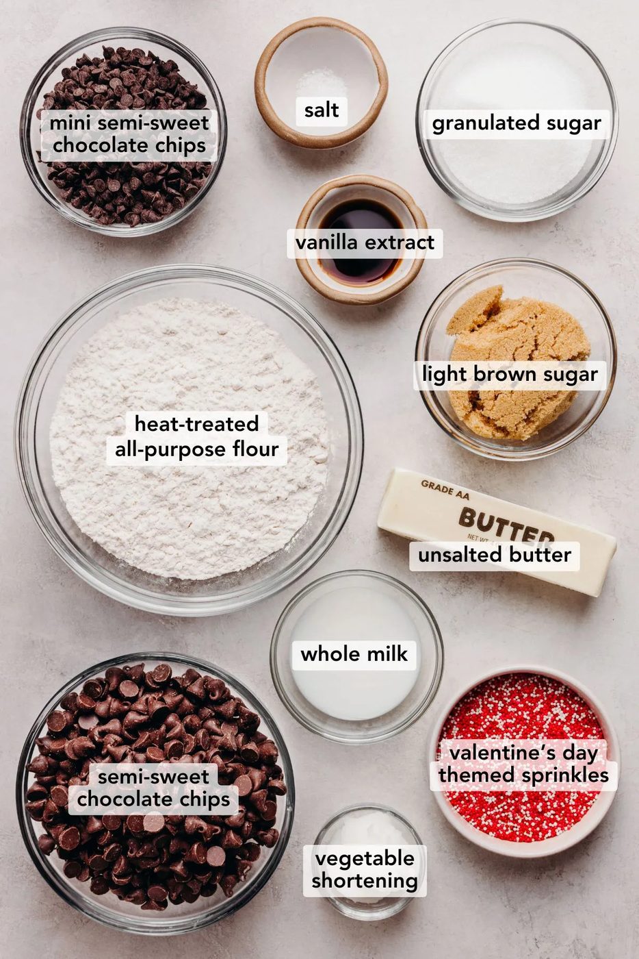 Ingredients in various size bowls labeled as all mini semi-sweet chocolate chips, salt, granulated sugar, light brown sugar, vanilla extract, heat treated all purpose flour, unsalted butter, whole milk, semi-sweet chocolate chips, vegetable shortening, and valentine's day themed sprinkles on a white marbled background.