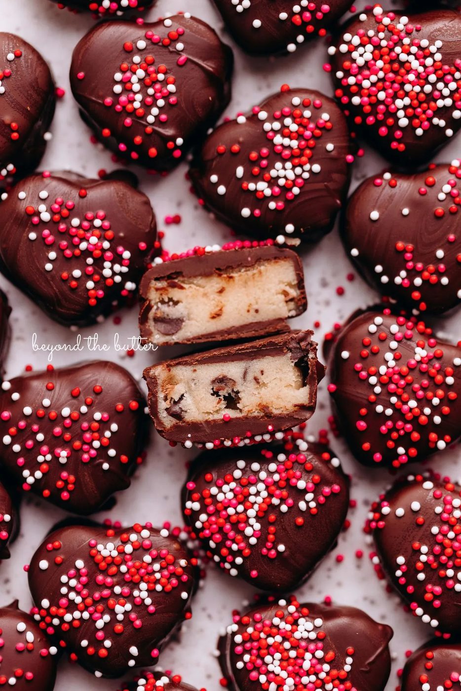 Chocolate covered cookie dough hearts topped with red, pink, and white sprinkles placed on a white parchment paper lined baking sheet with center heart cut in half to see the cookie dough inside.