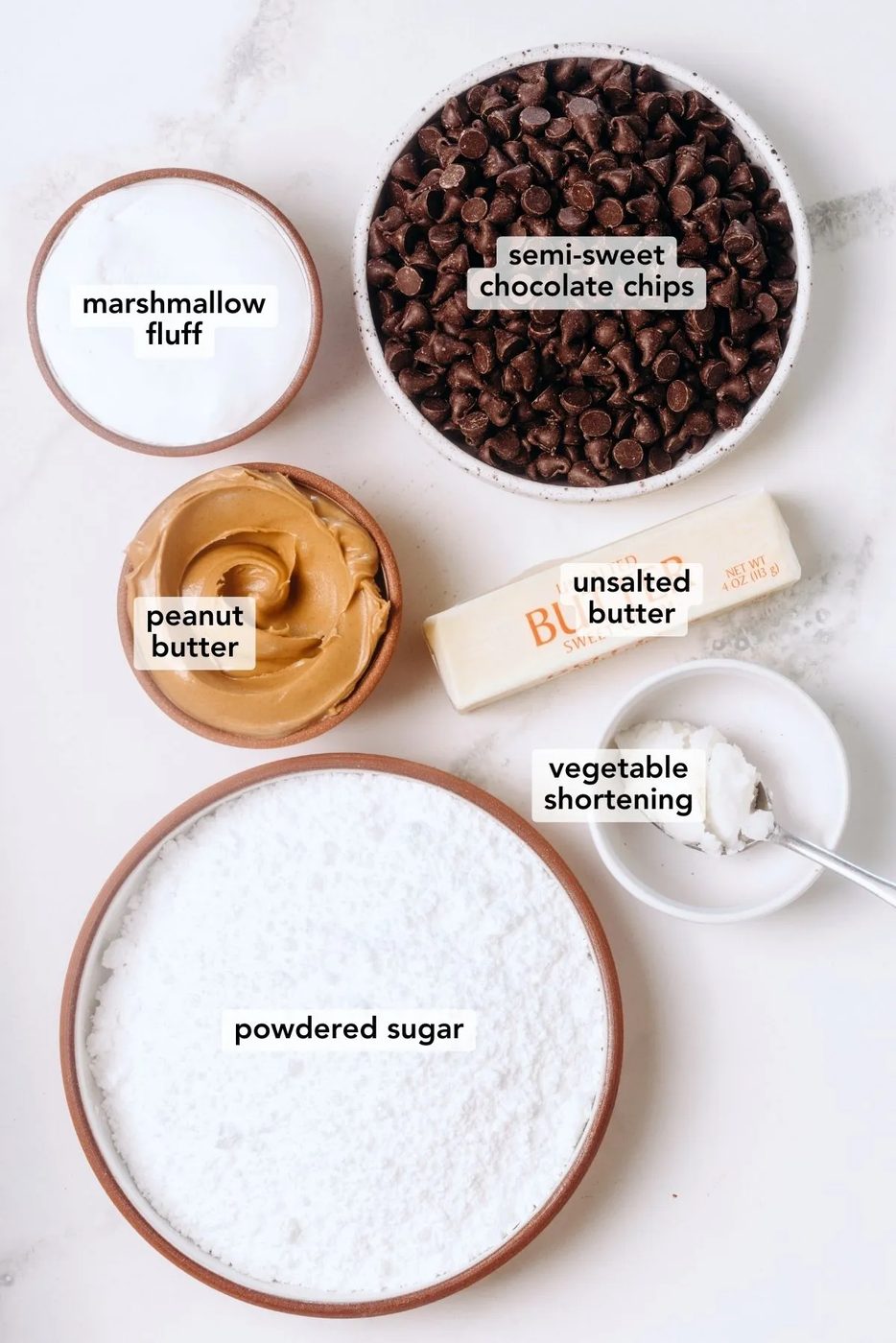 Ingredients in various size bowls labeled as semi-sweet chocolate chips, peanut butter, marshmallow fluff, unsalted butter, powdered sugar, and vegetable shortening on a white marbled background.
